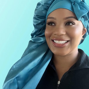 Puede incluir: Un gorro de dormir de sat&eacute;n azul turquesa con un lazo en la parte superior. El gorro lo lleva una persona de piel oscura y una gran sonrisa.