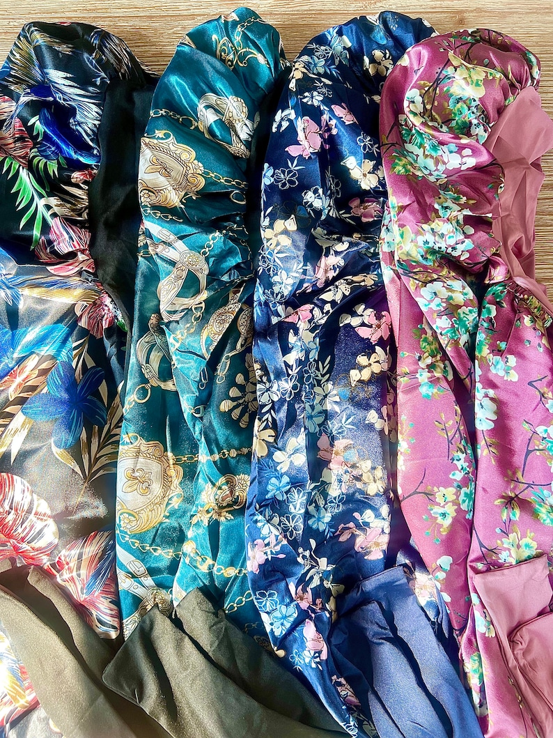 May include: Four satin hair bonnets in different patterns. The bonnets are teal with gold chains, navy blue with pink and white flowers, dark blue with white flowers, and burgundy with white flowers.