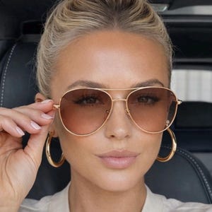 Può includere: Occhiali da sole aviator con montatura dorata e lenti sfumate marroni. Gli occhiali da sole sono tenuti leggermente sollevati. Sono visibili anche grandi orecchini a cerchio dorati. La persona ha i capelli biondi raccolti in uno chignon.