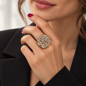 May include: A gold-toned statement ring featuring a large, circular cluster of clear, round-cut faceted crystals of varying sizes. The crystals are set closely together in a dense, circular arrangement, creating a brilliant, reflective surface.