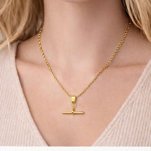 Peut inclure: Un collier en acier inoxydable plaqué or avec un pendentif en forme de barre en T. Le collier est présenté sur le cou d'une personne. Le texte sur l'image indique "18k PVD Gold Plated Stainless steel Waterproof • Tarnish Resistant."