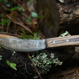 Puede incluir: Un cuchillo rústico hecho a mano con mango de madera y hoja de metal. El mango tiene tres remaches de metal. El cuchillo está apoyado en un trozo de madera.