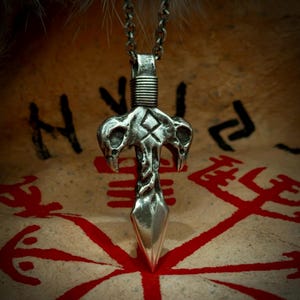 May include: A silver-colored pendant necklace featuring a stylized design with three skull-like shapes and a pointed bottom. The pendant hangs from a chain. The background shows a tan surface with red symbols.