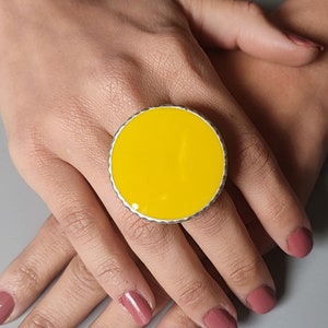 Puede incluir: Un anillo grande, redondo y amarillo con un borde plateado. El anillo se lleva en un dedo.