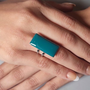 Könnte beinhalten: Ein rechteckiger Ring in blaugrüner Farbe mit einem silbernen Band. Der Ring befindet sich an einem Finger.