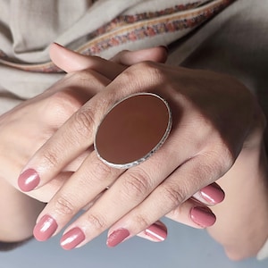 Könnte beinhalten: Ein großer ovaler Ring mit einem braunen Stein, der in Silber gefasst ist. Der Ring wird am Zeigefinger einer Hand mit rosafarbenem Nagellack getragen.