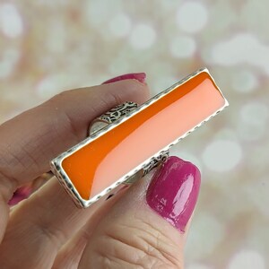 May include: A silver ring with a rectangular orange stone. The stone is set in a silver bezel and the ring band is decorated with a filigree design.