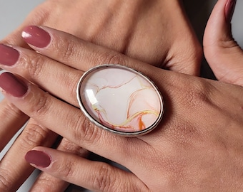 Può includere: Un anello d'argento con una grande pietra di resina di forma ovale. La pietra ha uno sfondo bianco con venature rosa e arancioni.