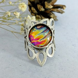 May include: A silver ring featuring a colourful abstract design in the centre. The ring has a decorative, openwork design around the centre piece.
