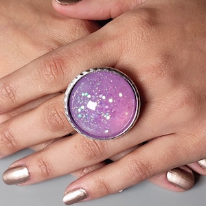 Pode incluir: Um anel grande e redondo com uma banda prateada. O anel apresenta um centro roxo claro cheio de glitter. O glitter é uma mistura de cores brancas e iridescentes. O anel é mostrado numa mão com unhas pintadas.