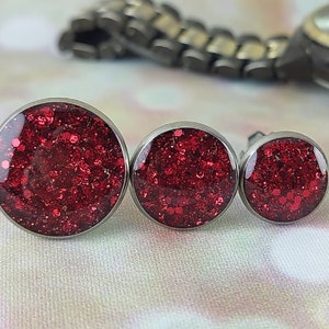 May include: Three round red glitter earrings in different sizes. The earrings are made of stainless steel and have a silver finish.
