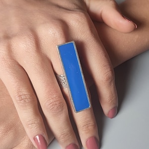 May include: A silver ring with a rectangular blue stone. The ring is worn on a finger.