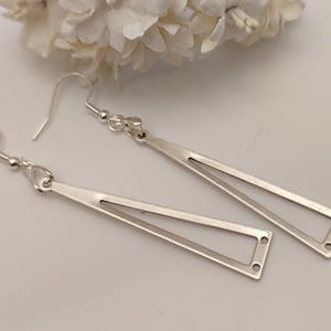 Könnte beinhalten: Ein Paar silberfarbene, geometrische Ohrhänger. Jeder Ohrring hat ein langes, offenes Dreieck-Design mit rechteckiger Basis. Die Ohrringe werden auf einer weißen Oberfläche mit weißen Blumen im Hintergrund präsentiert. Sie haben einen Hakenverschluss.