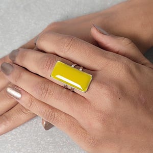 Könnte beinhalten: Ein Silberring mit einem leuchtend gelben, rechteckigen Stein. Der Ring hat ein modernes Design mit einem breiten Band und einem auffälligen gelben Stein. Der Ring wird an einer Hand mit polierten Nägeln gezeigt.