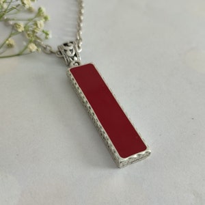 Puede incluir: Collar plateado con un colgante rectangular. El colgante presenta un centro rojo vibrante enmarcado por un borde detallado y estampado. La cadena es delicada y el diseño general es elegante.