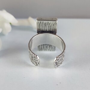 May include: A silver ring with a textured band and a square top. The top of the ring has a textured design.