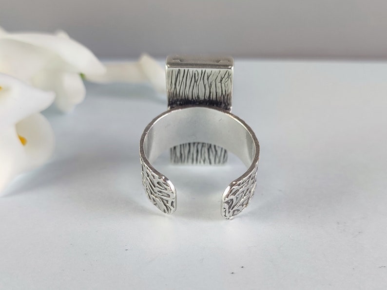May include: A silver ring with a textured band and a square top. The top of the ring has a textured design.