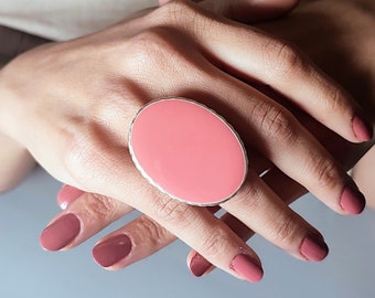 Può includere: Un anello grande di forma ovale con una pietra rosa incastonata in una fascia argentata. L'anello è indossato su un dito con smalto rosa.