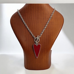 May include: A silver necklace featuring a red heart pendant. The heart-shaped pendant has a silver border. The necklace is displayed on a wooden bust. The chain is made of silver links.