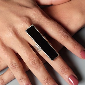 May include: A silver ring with a black rectangular stone. The ring is worn on the index finger of a hand.