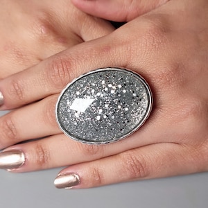 May include: A silver ring with a large oval-shaped stone. The stone is covered in silver glitter.
