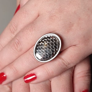 May include: A silver ring with a textured oval-shaped design that resembles fish scales. The ring is on a finger with red nail polish.