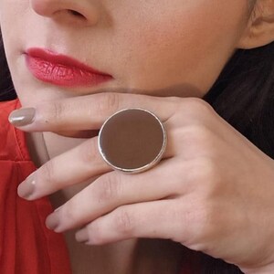 Pode incluir: Um anel grande e redondo com um centro castanho e uma banda prateada. O anel é uma peça marcante, com um design arrojado e minimalista. O anel é mostrado numa mão com batom vermelho e um top vermelho.
