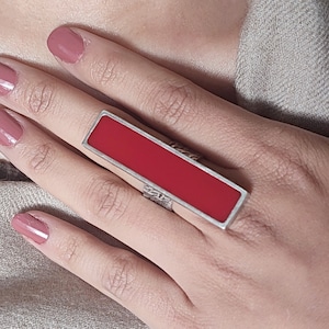 May include: A silver ring with a rectangular red stone. The ring is on a person's finger.