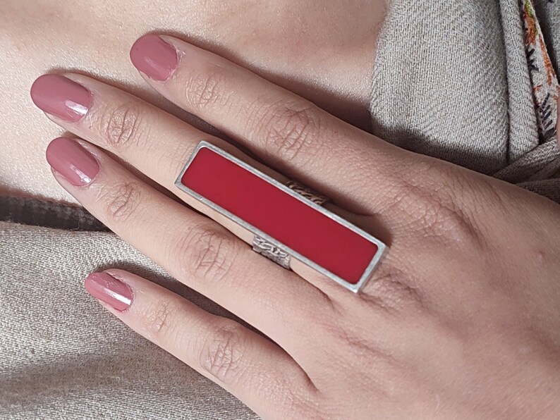 May include: A silver ring with a rectangular red stone. The ring is on a person's finger.