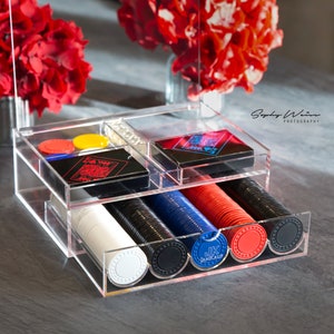 May include: A clear acrylic case with two drawers. The top drawer holds playing cards and the bottom drawer holds poker chips. The poker chips are red, blue, black, and white. The chips have the text "JK Jankauf" printed on them.