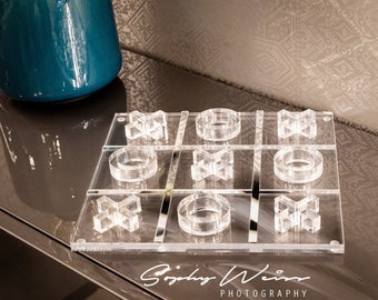 May include: A clear acrylic tic-tac-toe game board with a white background. The board has nine squares, each with a different symbol: a circle, an X, or a star. The board is sitting on a gray surface. The text "Sydney Weir Photography" is printed on the bottom of the board.