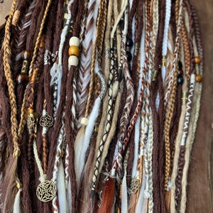 May include: A collection of brown, white, and beige synthetic dreadlocks with various beads and charms. The dreadlocks are arranged in a vertical row and are ready to be added to hair.