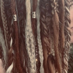 May include: Close-up of a person's hair styled in dreadlocks with various shades of brown and white. The dreadlocks are adorned with silver rings.