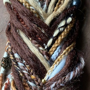 May include: A close-up of a braid made with brown, blonde, and white synthetic hair extensions. The braid is decorated with beads and metal charms.