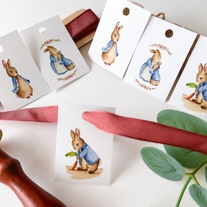 May include: Five white gift tags featuring watercolour illustrations of Peter Rabbit in various poses. The tags have a hole punched at the top and are tied with a red ribbon.