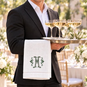 May include: A white linen towel featuring a dark green embroidered monogram letter M with decorative scrollwork, draped over the arm of a black suit jacket while holding a silver tray with four stemmed glasses filled with champagne.