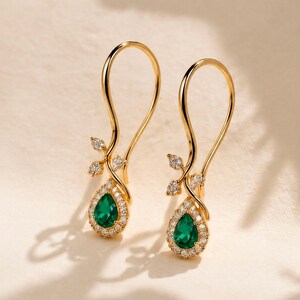Pode incluir: Um par de brincos pendentes de ouro com uma pedra preciosa verde em forma de pera rodeada por um halo de pequenas pedras brancas. Os brincos têm um design de fio curvo com um detalhe em forma de ramo adornado com três pequenas pedras brancas.