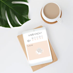 May include: A white tablet displaying a "Healthy Meal Daily Journal" layout, with mood trackers and a to-do list section. A cup of coffee sits on a cork coaster, and a large green leaf is visible. The tablet rests on a wooden board.