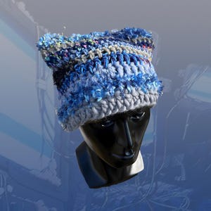 Puede incluir: Un gorro de ganchillo azul y gris hecho a mano con un diseño texturizado y esponjoso. El gorro presenta una mezcla de tonos azules y hilo gris, con un borde ligeramente elevado. El gorro se muestra en una cabeza de maniquí negra, mostrando su forma y estilo.