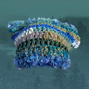May include: A handmade crochet hat in shades of blue, green, and grey. The hat features a textured design with a fluffy, fringed brim. The colour palette includes teal, navy, and olive green, creating a unique accessory.