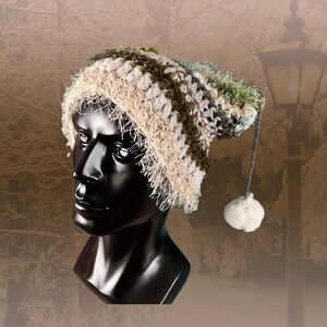 Pode incluir: Um chapéu de crochê feito à mão em tons neutros de bege, verde-oliva e cinza, com uma borda fofa. O chapéu apresenta uma longa corda cinza com um pompom branco na extremidade. O chapéu é exibido em uma cabeça de manequim preta.