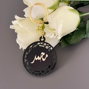 May include: Black and gold keychain with a circular pendant. The pendant features a gold inscription on a black background, surrounded by a decorative geometric border. The keychain has a gold ring and chain.