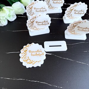 May include: White Ramadan Mubarak place card holders with gold calligraphy. The holders are scalloped-edged and feature a rectangular base. Several are arranged on a black and white marble-patterned surface, with white flowers in the background.