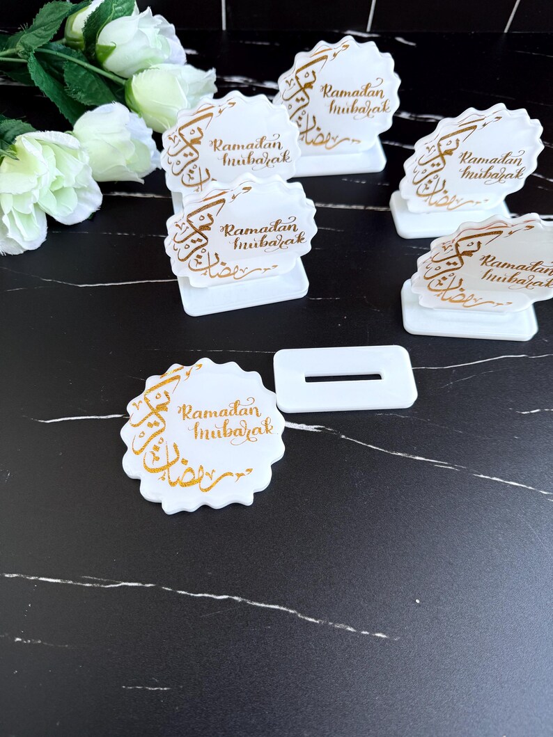 May include: White Ramadan Mubarak place card holders with gold calligraphy. The holders are scalloped-edged and feature a rectangular base. Several are arranged on a black and white marble-patterned surface, with white flowers in the background.