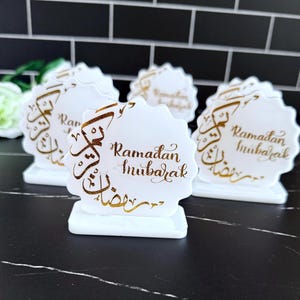 May include: White, scalloped-edge table decorations with gold calligraphy that reads "Ramadan Mubarak." The decorations are on a white base. The background is a black tile wall.