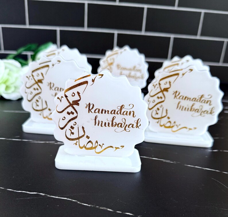May include: White, scalloped-edge table decorations with gold calligraphy that reads "Ramadan Mubarak." The decorations are on a white base. The background is a black tile wall.