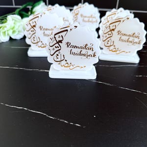 May include: White, scalloped-edge table decorations with gold script that reads "Ramadan Mubarak." The decorations are on a black surface. The decorations are in the shape of a flower.