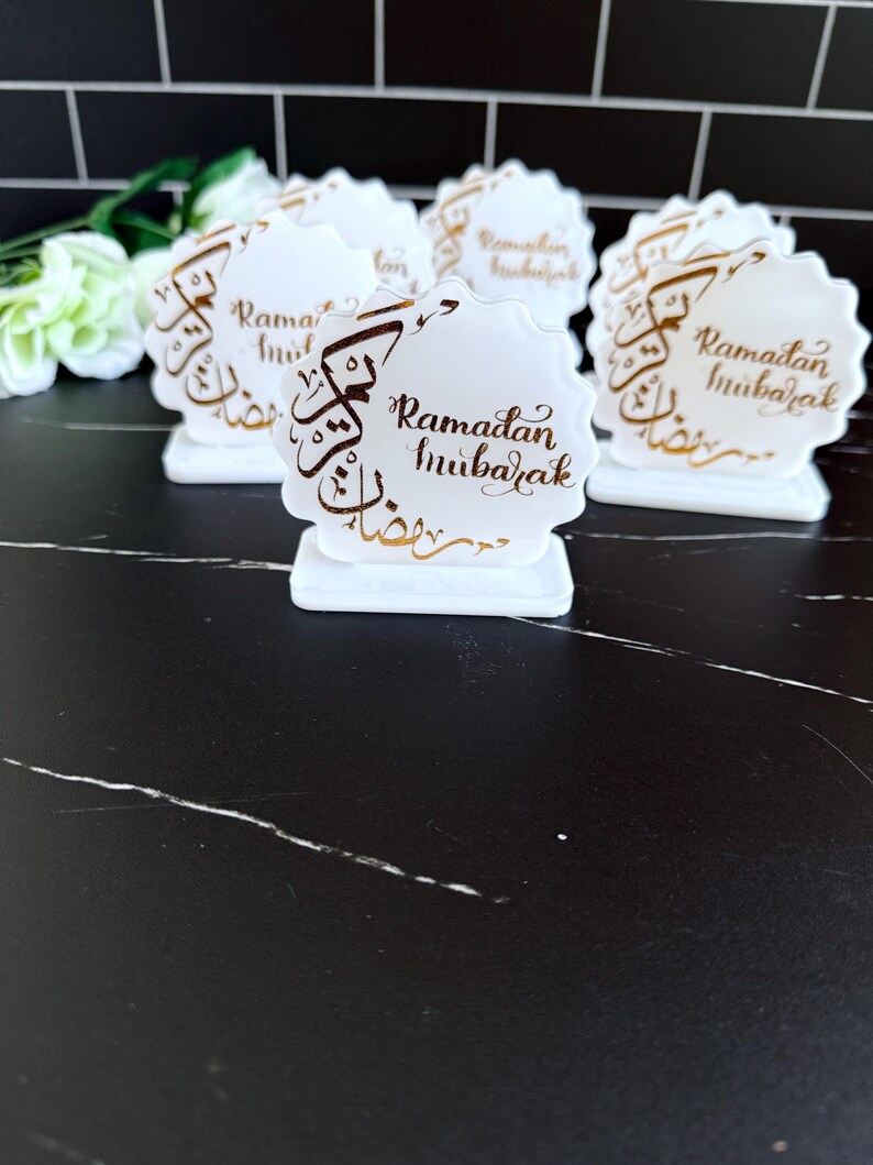 May include: White, scalloped-edge table decorations with gold script that reads "Ramadan Mubarak." The decorations are on a black surface. The decorations are in the shape of a flower.