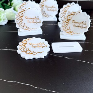 May include: White, scalloped-edge Ramadan Mubarak decorations with gold calligraphy. The decorations are on small white stands. One decoration is separate from its stand. The background is a black surface with white veining.