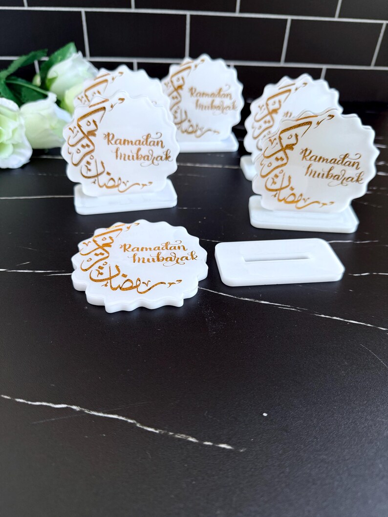May include: White, scalloped-edge Ramadan Mubarak decorations with gold calligraphy. The decorations are on small white stands. One decoration is separate from its stand. The background is a black surface with white veining.